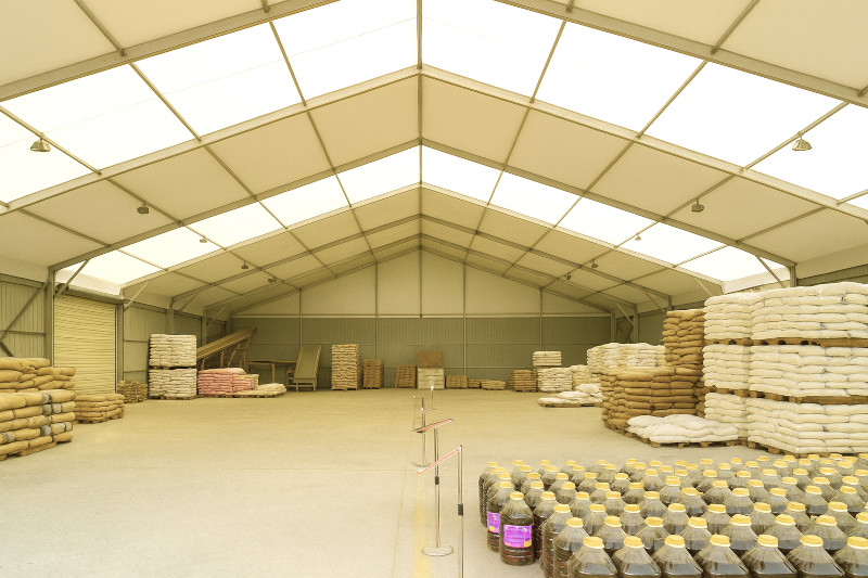 An interior view of a large storage facility with stacked bags of various materials, including grains. Containers with yellow caps are arranged in rows along the floor, under a high ceiling with ample natural light.