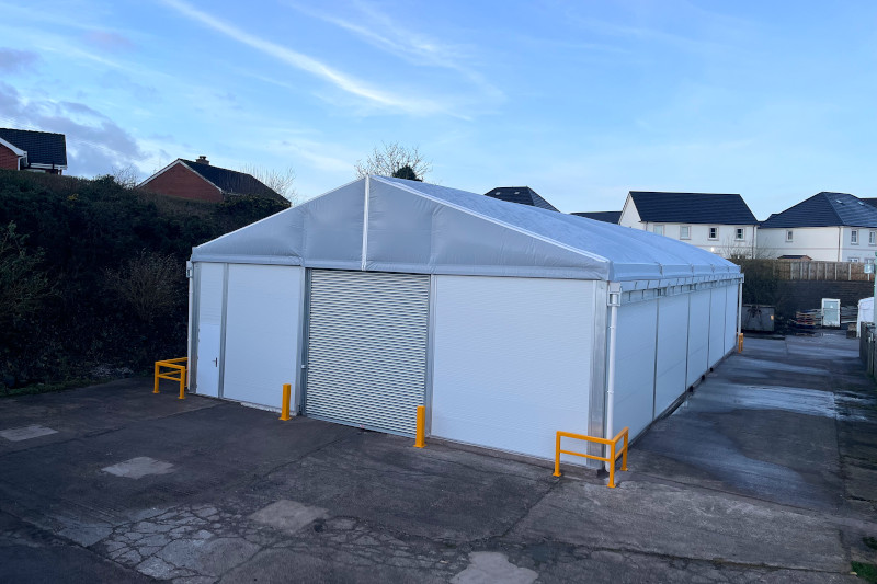 A large white fabric structure with a peaked roof, positioned on a concrete surface. The entrance features a roller shutter, with yellow barriers on either side. Residential buildings are visible in the background under a clear sky.